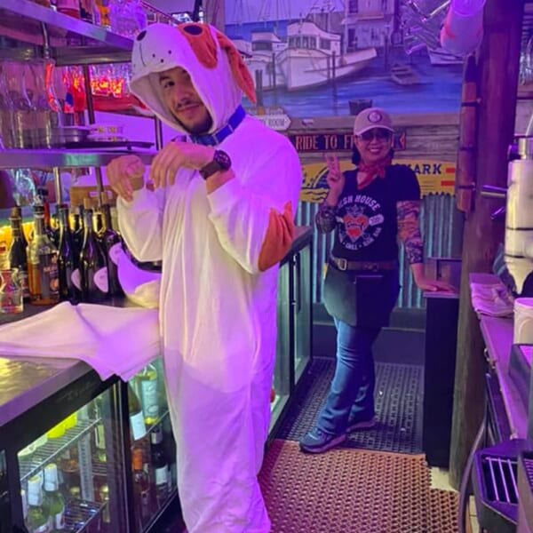 A man in a dog costume and a woman in a Fish House shirt pose behind a bar.