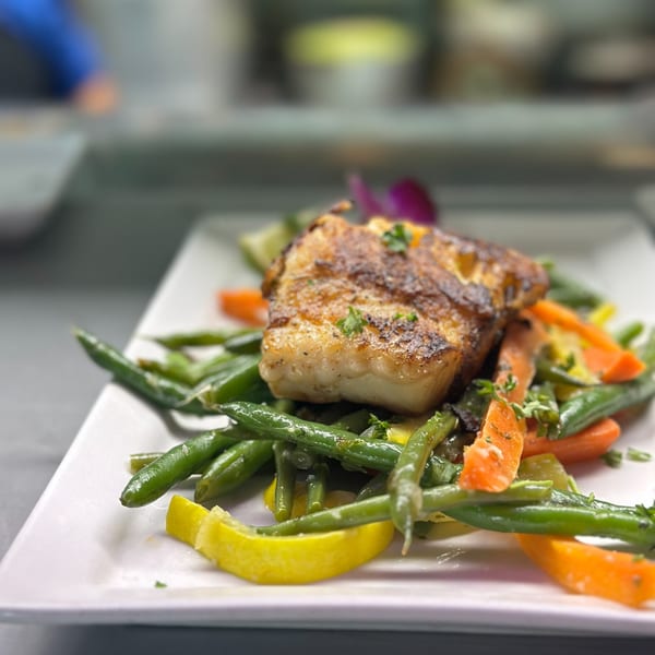 Grilled fish fillet with green beans, carrots, and lemon wedges on a white plate.