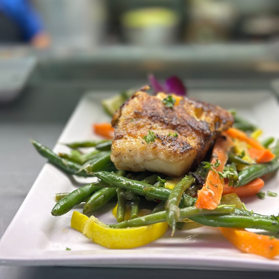 Grilled fish fillet with green beans, carrots, and lemon wedges on a white plate.