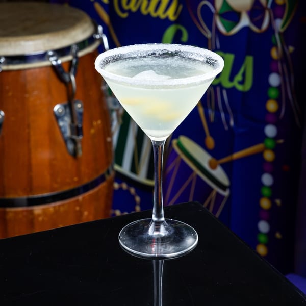 A martini glass with a salted rim and light-colored drink sits on a black surface, with a drum and Mardi Gras decorations in the background.