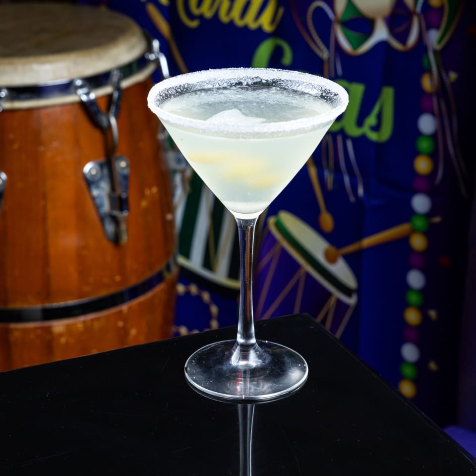 A martini glass with a salted rim and light-colored drink sits on a black surface, with a drum and Mardi Gras decorations in the background.