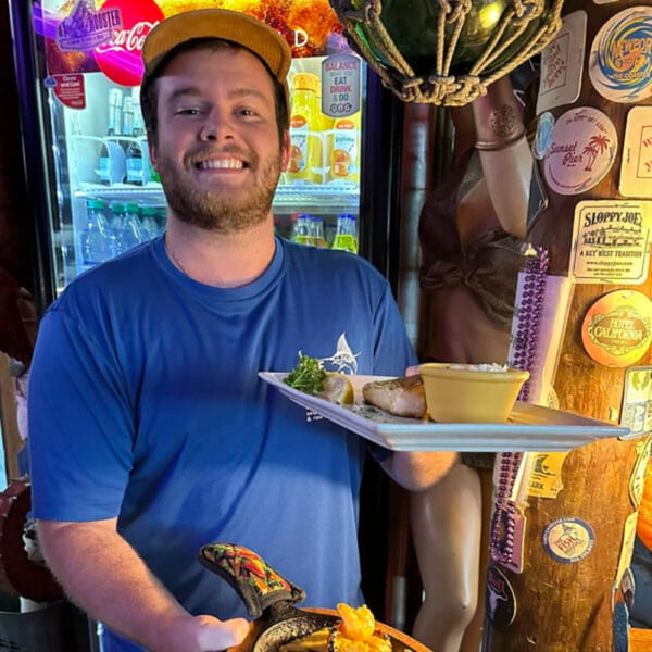 Smiling server in a blue shirt holding a plate of fish and a sizzling pan of shrimp.