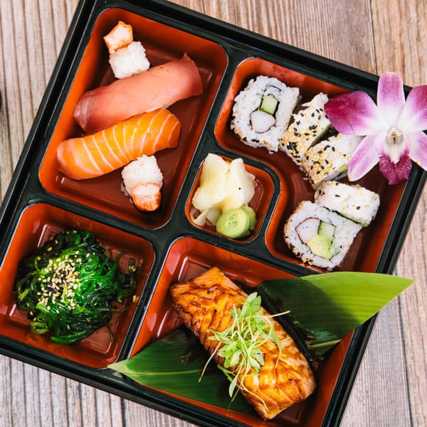 A bento box with sushi, salmon, seaweed salad, ginger, wasabi, and an orchid.