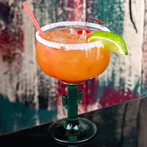 A vibrant orange cocktail in a cactus-stemmed glass, garnished with a lime wedge and cherry.