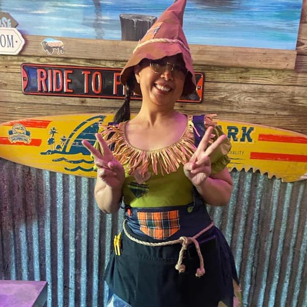 A woman in a scarecrow costume smiles and makes peace signs in front of a wooden wall.